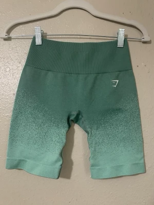 Gröna shorts Gymshark - Snygga gröna seamless träningsshorts från Gymshark med hög midja och coolt gradientmönster. Perfekta för gymmet eller löpning, med stretchigt material som sitter skönt och följer kroppen. Diskret logga på vänster sida.