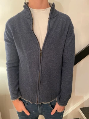 Blå kofta med dragkedja från LeeCooper - Säljer en blå kofta från LeeCooper med ribbad krage i grått och hel dragkedja framtill. Koftan är stickad och har långa ärmar samt ribbade muddar och nederkant. Perfekt för dig som gillar en chill och stilren look.