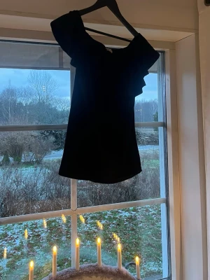 Svart offshoulder topp Gina Tricot - Säljer en svart offshoulder topp från Gina Tricot med volangdetalj över bröstet. Toppen har en mjuk och stretchig känsla, är ärmlös och har en snygg, feminin form. Passar M också 