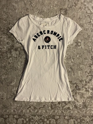Vit t-shirt Abercrombie & Fitch S - Vit vintage t-shirt från Abercrombie & Fitch i storlek S. Klassisk rund hals och korta ärmar. Tryck med logga och text på bröstet. Slimmad passform och mjukt bomullsmaterial som känns skönt mot huden. Perfekt för en chill och supersöt look. Inga defekter tillkommer! Dm för mått och frågor 💘