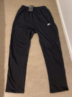Svarta mjukisbyxor från Nike - Svarta mjukisbyxor från Nike med elastisk midja och klassisk Nike-logga i vitt på vänster lår. Byxorna har en rak passform och är gjorda i mjukt bomullsmaterial, perfekta för chill eller träning. Snygg och enkel design med knappar i midjan för extra komfort.