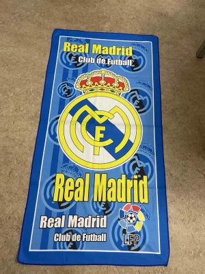 Real Madrid badhandduk i blått - Snygg badhandduk med Real Madrid-tryck i blått, gult och vitt. Handduken har klubbens stora logga i mitten, krona upptill och LFP-symbolen nedtill. Perfekt för fotbollsfans som vill visa sitt stöd på stranden eller vid poolen.
