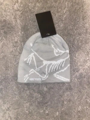 Grå mössa från Arc'teryx - Snygg grå mössa från Arc'teryx med vit logga och fossilmönster på framsidan. Mössan är stickad och har en mjuk, skön känsla som passar perfekt för kyliga dagar. Enkel design med diskret branding och stretchig passform.