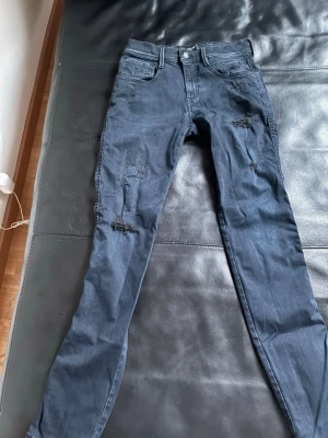 Replay Anbass mörkblå jeans med slitningar - Säljer ett par Replay Anbass jeans i mörkblå färg med snygga slitningar på benen. Modellen är slim och har klassisk femficksdesign. Jeansen är tillverkade i stretchigt bomullsmaterial för skön passform och har dragkedja och knapp framtill.