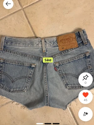 Levi's blå jeansshorts låg midja - Säljer ett par klassiska blå jeansshorts från Levi's med låg midja och råa, fransiga kanter. Shortsen har fem fickor, knappgylf och den ikoniska Levi's-lappen bak. Perfekta för varma dagar och chill stil.
