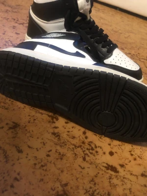 Nike Air Jordan 1 svart/vit sneakers - Säljer ett par Nike Air Jordan 1 sneakers i svart och vitt med klassisk high-top design. Skorna har snörning, perforerad tå och rund tå. Tillverkade i skinn med svart yttersula och vita detaljer. Märket syns tydligt på plösen och sidan. Perfekt för dig som gillar streetstyle.        OBS Kan gå ner i pris vid snabbafär.