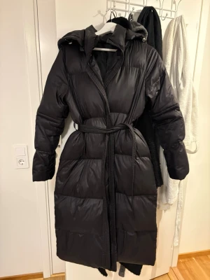 Svart lång pufferjacka med bälte - Säljer en svart lång pufferjacka med huva och bälte i midjan. Jackan har en quiltad design och är perfekt för kalla dagar. Den har långa ärmar och en oversized look som ger en riktigt skön vibe. Passar dig som vill hålla dig varm och snygg i vinter.