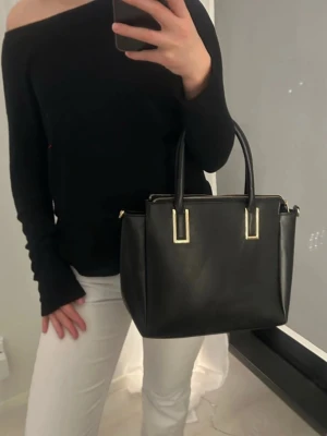 Svart handväska - Säljer en svart handväska med guldfärgade detaljer och dragkedja upptill. Väskan har två rejäla handtag och en rymlig insida med innerfack. Perfekt storlek för att få plats med allt nödvändigt och passar till många olika outfits🩷