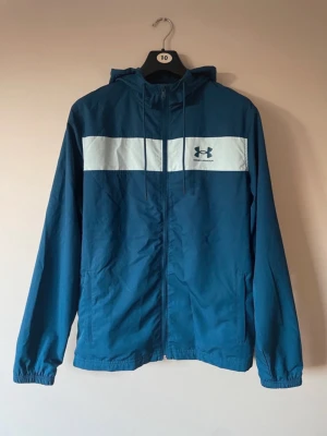 Blå Under Armour windbreaker - Snygg blå vindjacka från Under Armour med vit panel över bröstet och logga. Jackan har huva med snörning, dragkedja framtill och meshfoder på insidan. Perfekt för dig som gillar sportig stil och vill ha något lätt och funktionellt.
