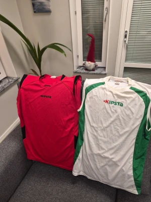 Kipsta Sporttröjor - 2 st - Storlek M - Två ärmlösa sporttröjor från Kipsta. En röd med svarta detaljer och en vit med gröna detaljer. Lite missfärgat, kolla bilerna.