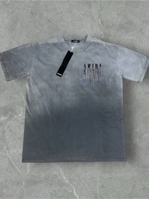 Grå t-shirt från Amiri med färgstänk - Säljer en grå t-shirt från Amiri med coolt färgstänksmönster och stor logga både fram och bak. T-shirten har en relaxed passform och är tillverkad i mjuk bomull. Perfekt för dig som gillar streetwear och vill sticka ut med snygga detaljer. Står ej vilken storlek men när jag köpte den stod det storlek M på hemsidan