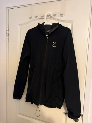 Svart vindjacka från Haglöfs - Svart vindjacka från Haglöfs med huva och hel dragkedja framtill. Jackan har två sidofickor med dragkedja och justerbar nederkant med snörning. Perfekt för utomhusaktiviteter och har en stilren look med diskret logga på bröstet.