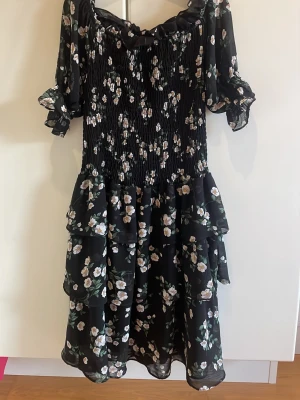 Blommig svart klänning från IVIVI - Supersöt svart klänning från IVIVI i storlek S med vita och ljusgula blommor. Klänningen har volanger, smockad överdel och korta puffärmar. Offshoulder-design och flowy kjol ger en romantisk vibe. Perfekt för dig som gillar detaljer och blommigt!