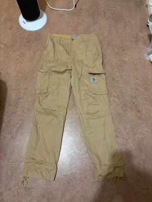 Beiga cargopants från Carhartt WIP - Säljer ett par beiga cargopants från Carhartt Work In Progress i storlek M. Byxorna har flera stora fickor med lock, snörning vid benslut och klassisk Carhartt-logga på benet. Tillverkade i slitstark bomull, perfekta för dig som gillar streetwear och funktion. Sitter perfekt som S