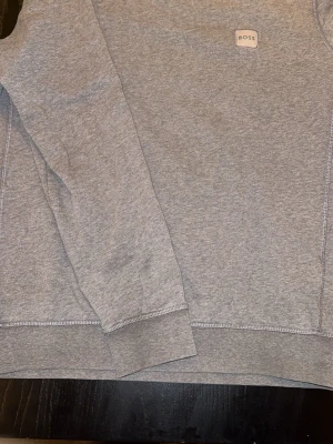Grå sweatshirt från BOSS - Säljer en stilren grå sweatshirt från BOSS med klassisk rund hals och ribbade muddar. Tröjan har ett litet BOSS-märke på bröstet och är tillverkad i mjuk bomull. Perfekt för chill dagar och enkel att matcha med jeans eller joggers.