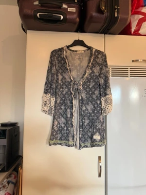 Mönstrad kofta från Odd Molly - Säljer en unik kofta från Odd Molly med blått och vitt mönster, kontrasterande ärmslut och gröna volangdetaljer längst ner. Koftan har trekvartsärm, v-ringning och knyt framtill. Perfekt för dig som gillar boho-vibe och snygga detaljer. Storlek 1 som enligt deras storleksguide motsvarar en storlek S. Mycket fint skick, jag hittar inga synliga anmärkningar eller defekter. 