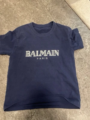 Mörkblå Balmain Paris t-shirt - Snygg mörkblå t-shirt från Balmain Paris med vit logotyp på bröstet. Klassisk rund hals och korta ärmar. Tillverkad i mjuk bomull som känns skön mot huden. Perfekt för dig som gillar stilrena designer och vill ha ett statement-plagg i garderoben.