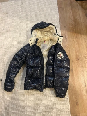 Moncler pufferjacka med huva - Säljer en pufferjacka från Moncler med glansig finish och beige insida. Jackan har huva, dragkedja framtill och ett stort Moncler-märke på bröstet. Flera praktiska fickor med dragkedja och snygga detaljer. Perfekt för kalla dagar. XS (0)