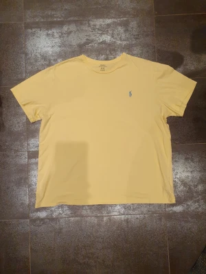 Gul t-shirt från Polo Ralph Lauren XL - Snygg gul t-shirt från Polo Ralph Lauren i storlek XL. Klassisk design med rund hals och korta ärmar, samt den ikoniska blå Polo-loggan broderad på bröstet. Tillverkad i mjuk bomull som känns skön mot huden. Perfekt för dig som gillar stilrena och enkla plagg.