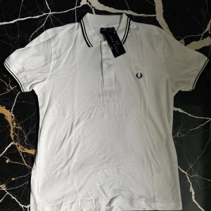 Vit polotopp från Fred Perry - Säljer en klassisk vit polotopp från Fred Perry med svart broderad logga på bröstet. Toppen har korta ärmar, krage med svarta ränder och knappar framtill. Materialet är mjuk bomull och passformen är normal. Perfekt för en clean och sportig look.