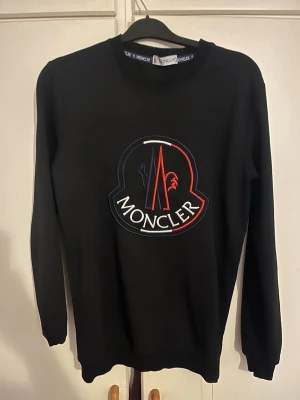 Svart sweatshirt från Moncler - Säljer en svart sweatshirt från Moncler med stort broderat logotyptryck i vitt, rött och blått på bröstet. Tröjan har rund hals och långa ärmar med ribbade muddar. Perfekt för dig som gillar stilrena märkesplagg.