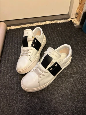 Valentino Garavani vita sneakers - Säljer ett par snygga Valentino Garavani sneakers i vitt skinn med bred svart detalj över snörningen. Skorna har platt sula med mönstrad undersida och rund tå. Guldtext på plösen och coola nitar längs hälen. Perfekt för dig som gillar exklusiva märken och stilren design. Riktigt grisch!