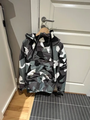Camouflage vindjacka från Dope XS - Säljer en fet skidjacka från Dope i storlek XS med grå, svart och vit camouflage-mönster. Jackan har huva med snörning, flera dragkedjor och stora fickor framtill. Perfekt för dig som gillar streetstyle och vill sticka ut med cool design. Perfekt till vintersäsongen!