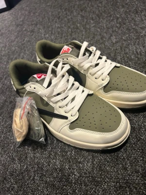Nike Air Jordan 1 Low Travis Scott - Säljer ett par Nike Air Jordan 1 Low Travis Scott med olivgröna och vita paneler, svart swoosh och röda broderade detaljer på hälen. Skorna har platt sula, rund tå och snörning. Materialet är skinn och syntet. Extra skosnören medföljer. Perfekt för dig som gillar streetwear.