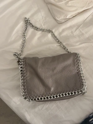 Grå axelväska med kedja - Säljer en snygg grå axelväska med grov silverkedja som axelrem och detaljer längs kanterna. Använd några gånger 🥰