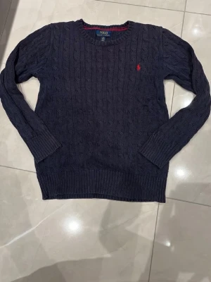 Mörkblå stickad tröja Polo Ralph Lauren - Säljer en mörkblå stickad tröja från Polo Ralph Lauren med klassiskt kabelstickat mönster och röd broderad logga på bröstet. Tröjan har rund hals och långa ärmar med ribbade muddar. Perfekt för dig som gillar stilren och tidlös design.
