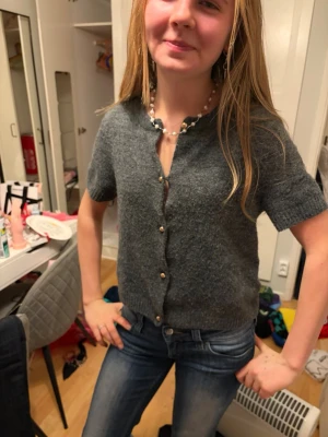 Grå stickad kofta med kort ärm - Mysig grå stickad kofta med korta ärmar och snygga guldfärgade knappar framtill. Perfekt att slänga på sig över ett linne eller t-shirt. Koftan har en klassisk passform och ribbade kanter vid ärmslut och nederkant.