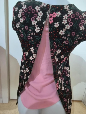 Blommig blus med dragkedja - Säljer en blommig blus med svart botten och mönster i rosa, vitt och grönt. Blusen har korta ärmar och en lång dragkedja som går längs hela ryggen, vilket ger en cool detalj. Materialet känns mjukt och lätt, perfekt för vår och sommar.