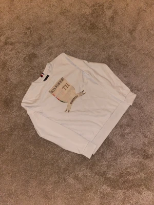 Vit sweatshirt från Tommy Hilfiger - Säljer en vit sweatshirt från Tommy Hilfiger med stort tryck på bröstet i form av ett sköldmotiv med texten 'HILFIGER TH' och romerska siffror. Tröjan har rund hals och långa ärmar, perfekt för en chill och stilren look.