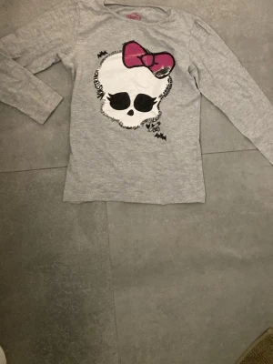 Monster High långärmad topp grå - Säljer en grå långärmad topp från Monster High med tryck av en vit döskalle med svart ögon och en stor rosa rosett. Toppen är i mjuk bomull och har rund hals. Perfekt för dig som gillar coola prints och Monster High.