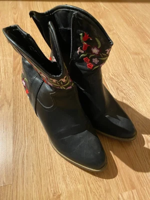 Svarta cowboy boots med blommig brodyr - Säljer ett par svarta cowboy boots med snygg spetsig tå och klack. Bootsens skaft är dekorerade med broderade blommor i rött, lila och grönt som ger en unik vibe. Tillverkade i skinn med dragkedja på insidan för enkel på- och avtagning.