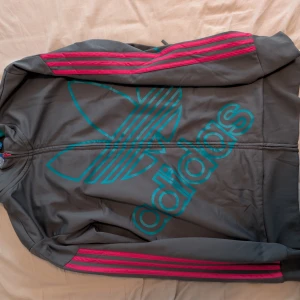 Grå Adidaströja med rosa detaljer - Snygg grå tröja från Adidas med stor turkos logga på framsidan och rosa ränder längs ärmarna. Jackan har dragkedja framtill och långärmade armar. Katt finns i hemmet. 