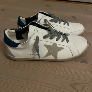 Golden Goose vita sneakers med stjärna - Golden Goose sneakers i vitt skinn med grå mockastjärna på sidan och grå snörning. Hälen har en blå detalj och sulan är platt och beige. Snygg, rund tå och klassisk low-top modell som ger en cool vibe till din outfit.