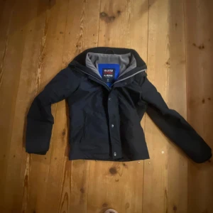 Svart vindjacka Hollister S - Svart vindjacka från Hollister med broderad logga på ryggen och mjukt grått foder. Jackan har dragkedja och knappar framtill, ribbad krage och långärmade ärmar. Perfekt för dig som gillar stilren och sportig look.