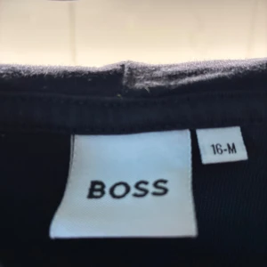 Svart hoodie från BOSS med guldtryck - Svart hoodie från BOSS i storlek M med guldiga detaljer på ärmarna och stort BOSS-tryck på bröstet. Tröjan har huva, lång ärm och en stor magficka framtill. Perfekt för dig som gillar stilrena streetwear-plagg med snygga detaljer.