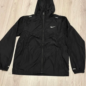 Svart Nike vindjacka med huva - Svart vindjacka från Nike med huva och hel dragkedja framtill. Jackan har långa ärmar, elastiska muddar och reflekterande detaljer. Materialet är lätt och syntetiskt, perfekt för blåsiga dagar. Swoosh-logga på bröstet ger en sportig vibe.