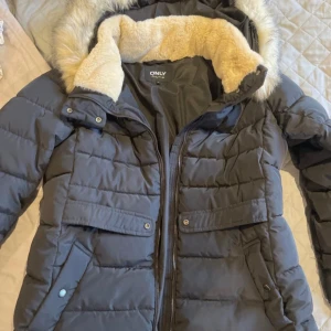 Svart pufferjacka med fuskpäls från ONLY - Säljer en svart pufferjacka från ONLY med fluffig fuskpäls runt luvan och quiltad design. Jackan har dragkedja, knappar och stora fickor framtill. Insidan av luvan är mjuk och beige, perfekt för kalla dagar. Snygg och praktisk vinterjacka! Knappt använd!