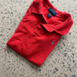 Röd pikétröja från Polo Ralph Lauren - Snygg röd pikétröja från Polo Ralph Lauren med klassisk krage och två knappar framtill. Tröjan har det ikoniska blå broderade Polo-logot på bröstet och är tillverkad i 100% bomull för en skön och luftig känsla. Perfekt för dig som gillar stilrena och sportiga plagg. Piken har blivit sydd vid magen kolla bild 5! 10-12 år