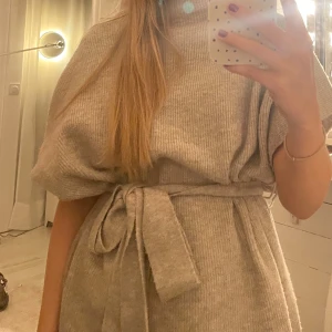 Beige stickad klänning med bälte - Supermysig beige stickad klänning med korta ärmar och högt hals. Klänningen har ett matchande bälte i midjan som knyts för en snygg siluett. Perfekt för dig som gillar en chill och trendig look.