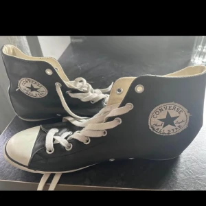 Svarta Converse All Star high-tops - Klassiska svarta Converse All Star high-top sneakers med vit tå och vita snören i skinn. Skorna har den ikoniska Converse-loggan på sidan och en platt sula. Perfekta för en cool och avslappnad stil.