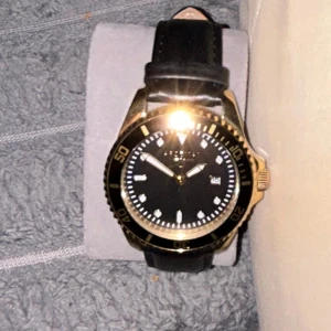 Arsenal klocka  - Stilren klocka från Arsenal  med svart urtavla, guldig boett och svarta detaljer. Klockan har datumvisning och svart läderarmband. Perfekt accessoar för dig som gillar klassisk och elegant design. Kommer med original box och pris kan diskuteras. 