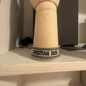 Christian Dior J'Adior armband - Snyggt armband från Christian Dior med texten 'J'Adior' broderad i svart på en vit bakgrund. Armbandet har en bred, vävd design med svarta detaljer och avslutas med två stilrena tofsar. Perfekt för dig som vill ha en lyxig och trendig accessoar.