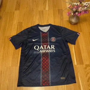 PSG matchtröja Nike Qatar Airways - Snygg PSG fotbollströja från Nike med Qatar Airways-tryck på bröstet. Tröjan är marinblå med rött och vitt grafiskt mönster längs mitten, broderad Nike-logga och PSG-emblem. Tillverkad i lätt och ventilerande polyester, perfekt för match eller träning.