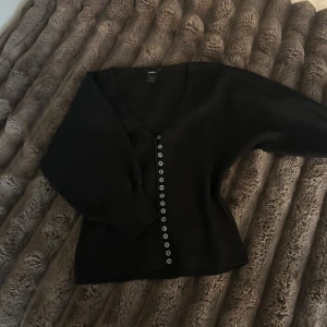 Svart kofta med knappar  - Svart kofta från Lindex med v-ringning och långa utsvängda  ärmar. Koftan har en rad med silvriga knappar framtill och är i ett mjukt, stickat material. Figursydd 