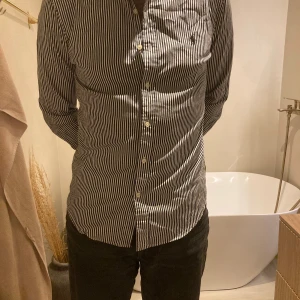 Randig skjorta från Ralph Lauren M - Slim fit skjorta från Ralph Lauren i svart och vitt med vertikala ränder. Klassisk krage, knappar framtill och broderad logga på bröstet. Tillverkad i mjuk bomull, perfekt för dig som gillar stilrena och snygga detaljer.
