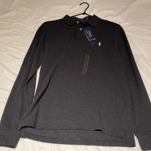 Mörkgrå pikétröja Polo Ralph Lauren - Snygg mörkgrå långärmad pikétröja från Polo Ralph Lauren med klassisk krage, två knappar och ikonisk broderad logga på bröstet. Modellen är custom slim fit och har en stilren look som funkar till många outfits. Materialet är mjuk bomull. Aldrig använd